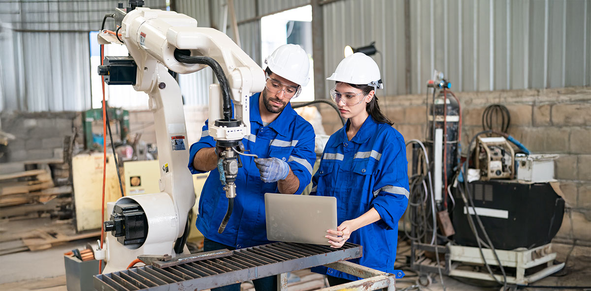 Industrial engineers in blue uniforms and safety gear operating a robotic arm in a manufacturing facility, illustrating leadership and workforce adaptation in the Industry 4.0 era.