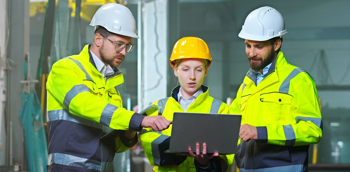 Team of engineers in safety gear analyzing data on a laptop inside a factory, demonstrating teamwork and process optimization in Manufacturing Partner Programs.